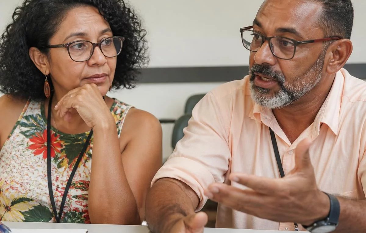Duas pessoas sentadas de frente uma para a outra à mesa: uma fala, a outra registra por escrito